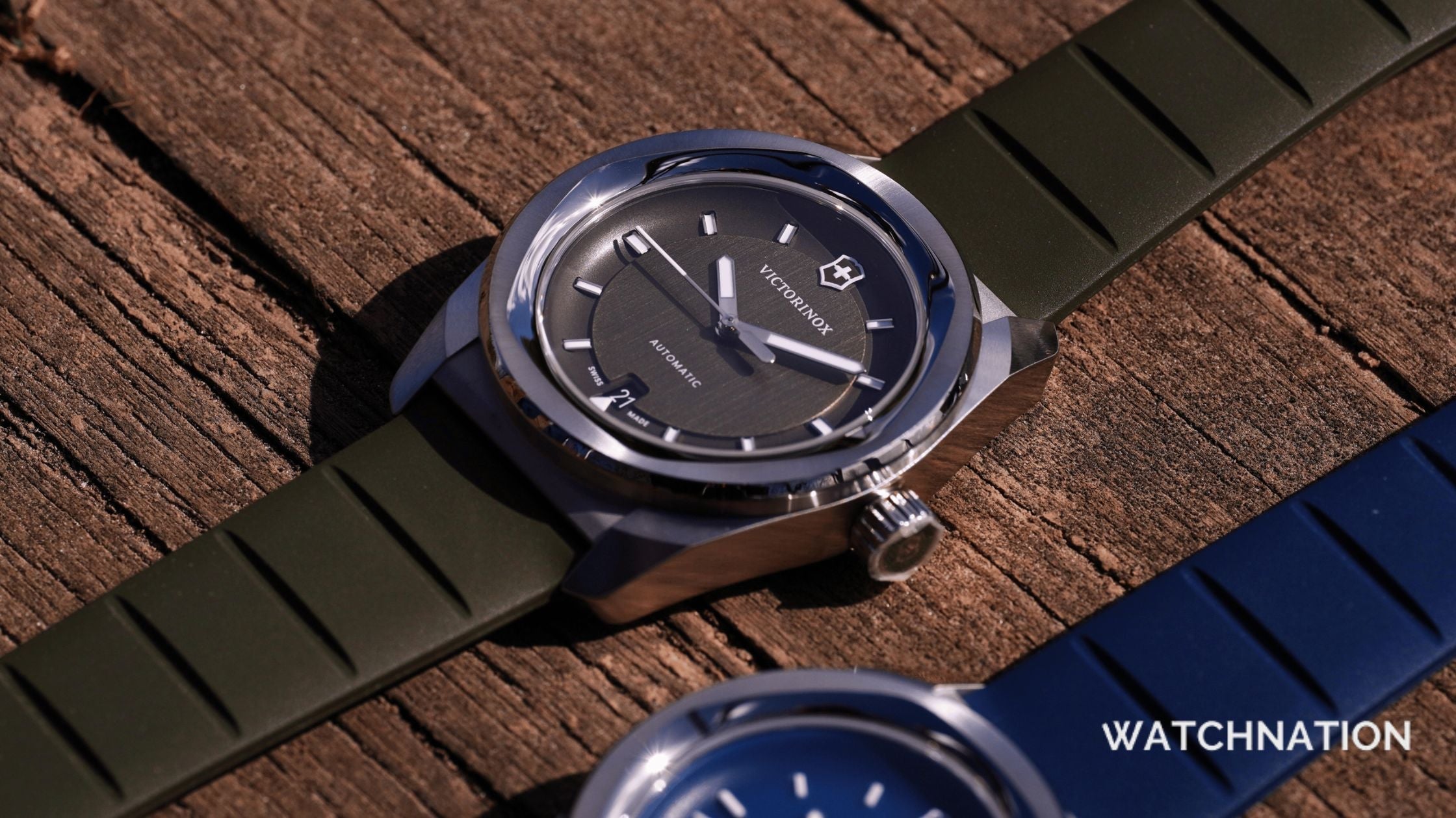 A close-up shot of a Victorinox automatic watch with a dark grey dial and an olive green rubber strap, resting on a textured wooden surface next to a blue strap version.