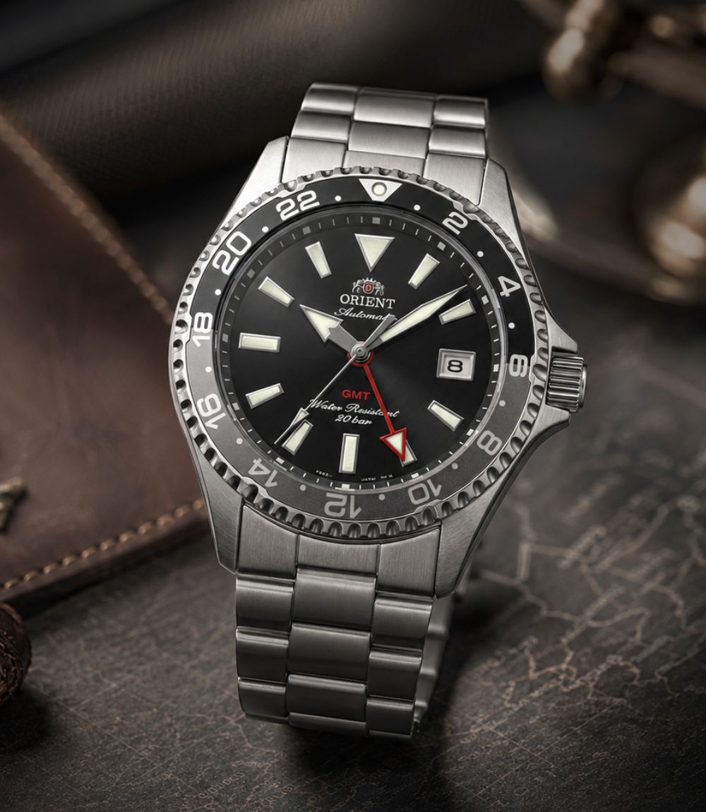 Orient Mako GMT automatic watch with a black dial, red GMT hand, and stainless steel bracelet resting on a dark leather background