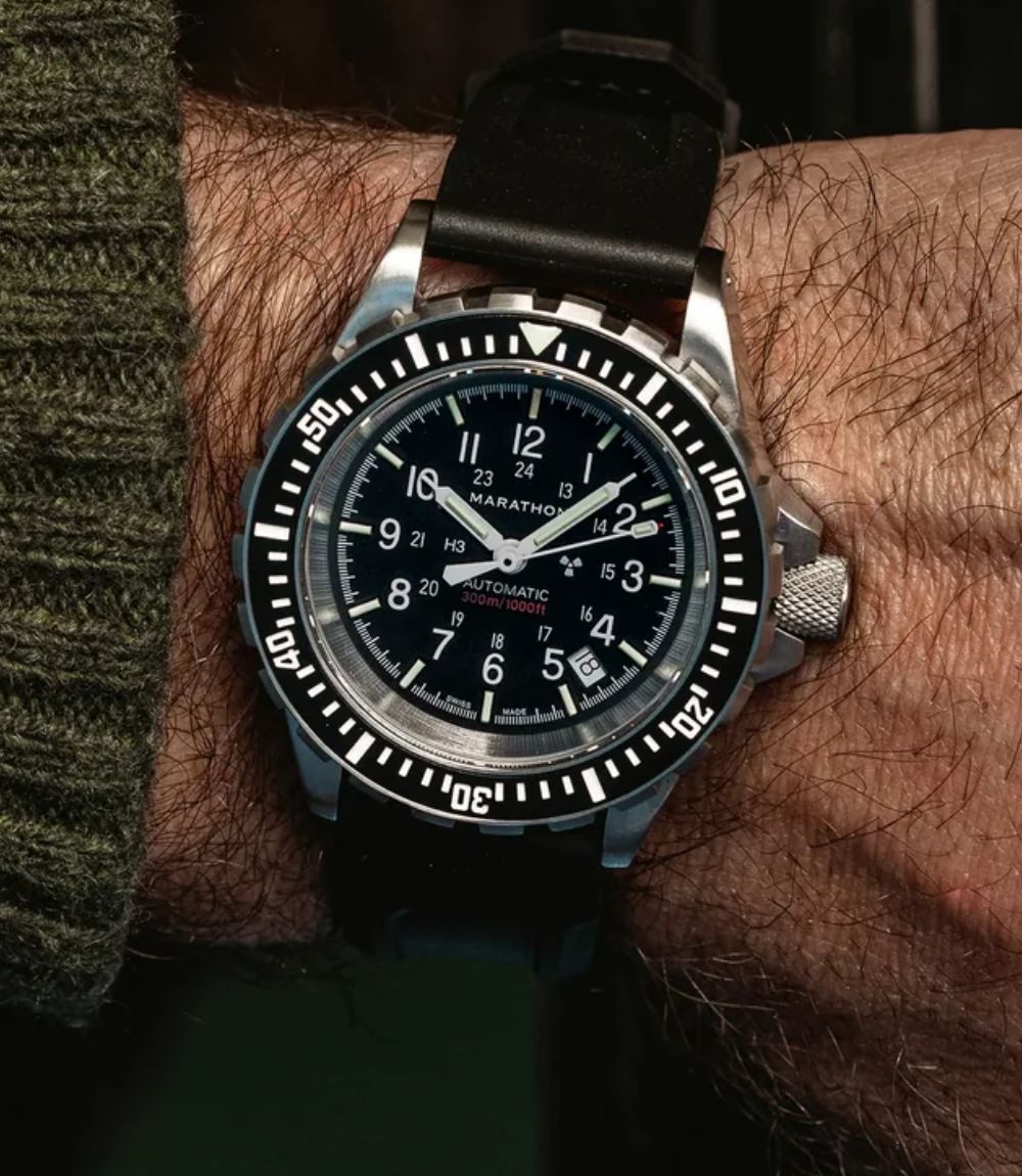 A close-up of a wrist wearing a Marathon black-dial dive watch with a black rubber strap and a silver-toned case