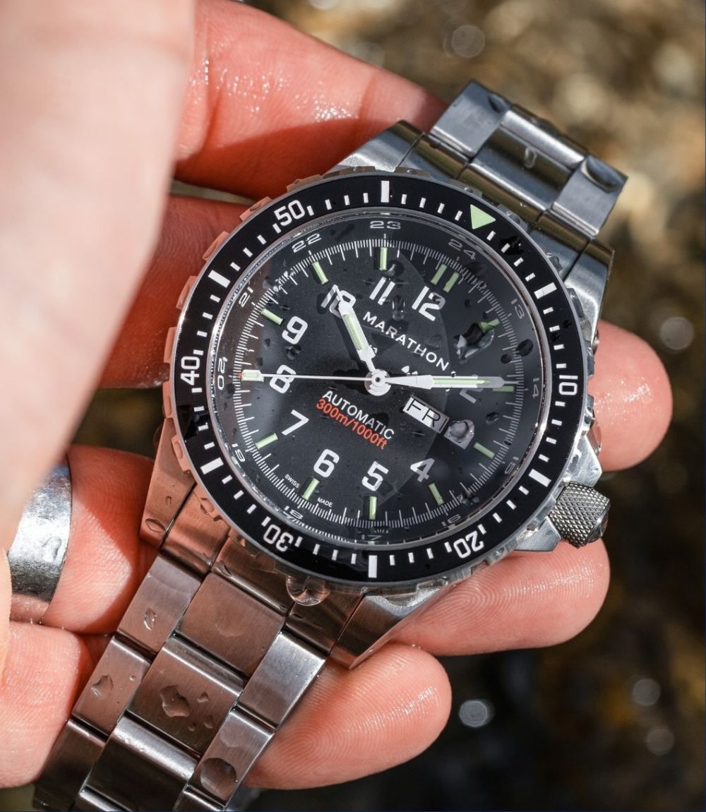 Marathon automatic watch with stainless steel bracelet held in hand against an outdoor background