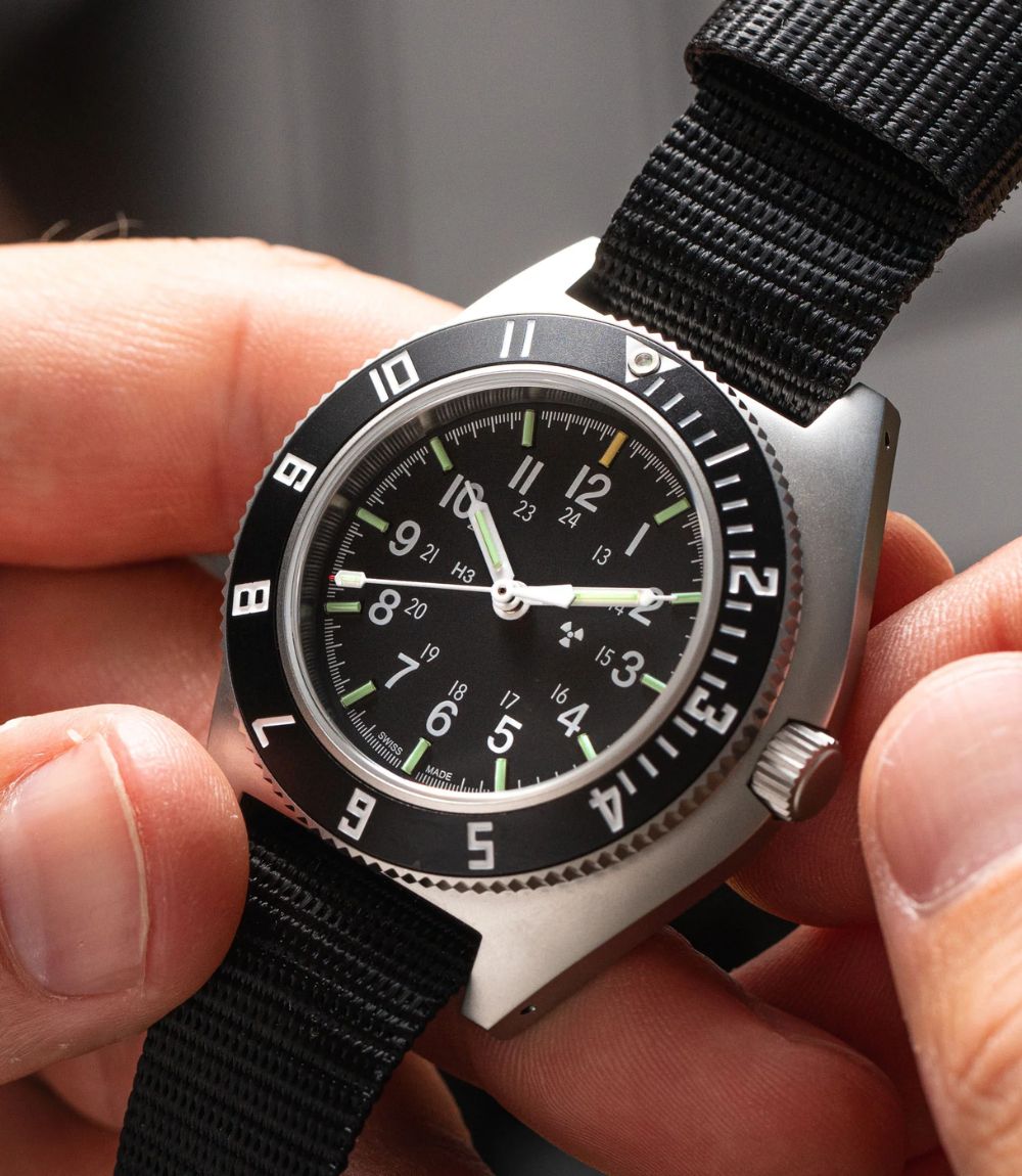 A close-up of a Marathon wristwatch being held by two hands. The watch has a black dial with luminous green and white numerals and hands, a black rotating bezel, and a silver-toned case with a black nylon strap