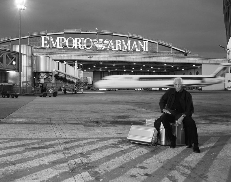 Black and white photograph of a man sitting on a suitcase on an airport tarmac in front of a large Emporio Armani terminal building, with a blurred airplane passing by in the background