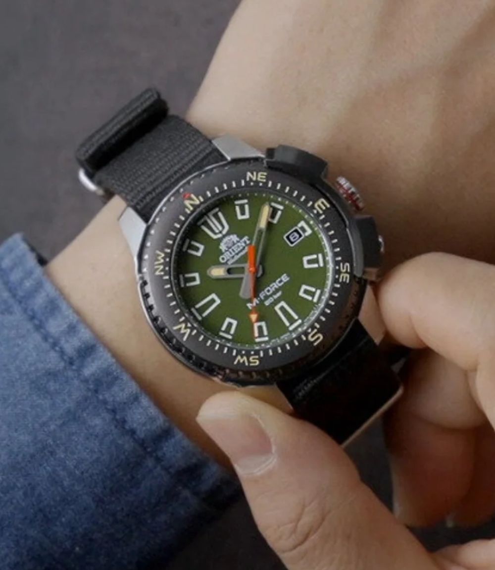Orient M-Force watch displaying a green dial and compass bezel worn on a wrist with a black fabric strap