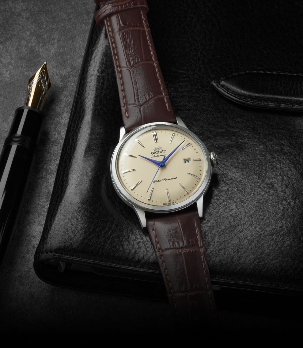 Orient Bambino dress watch featuring a cream dial, blue hands, and dark brown leather strap resting on a black leather journal