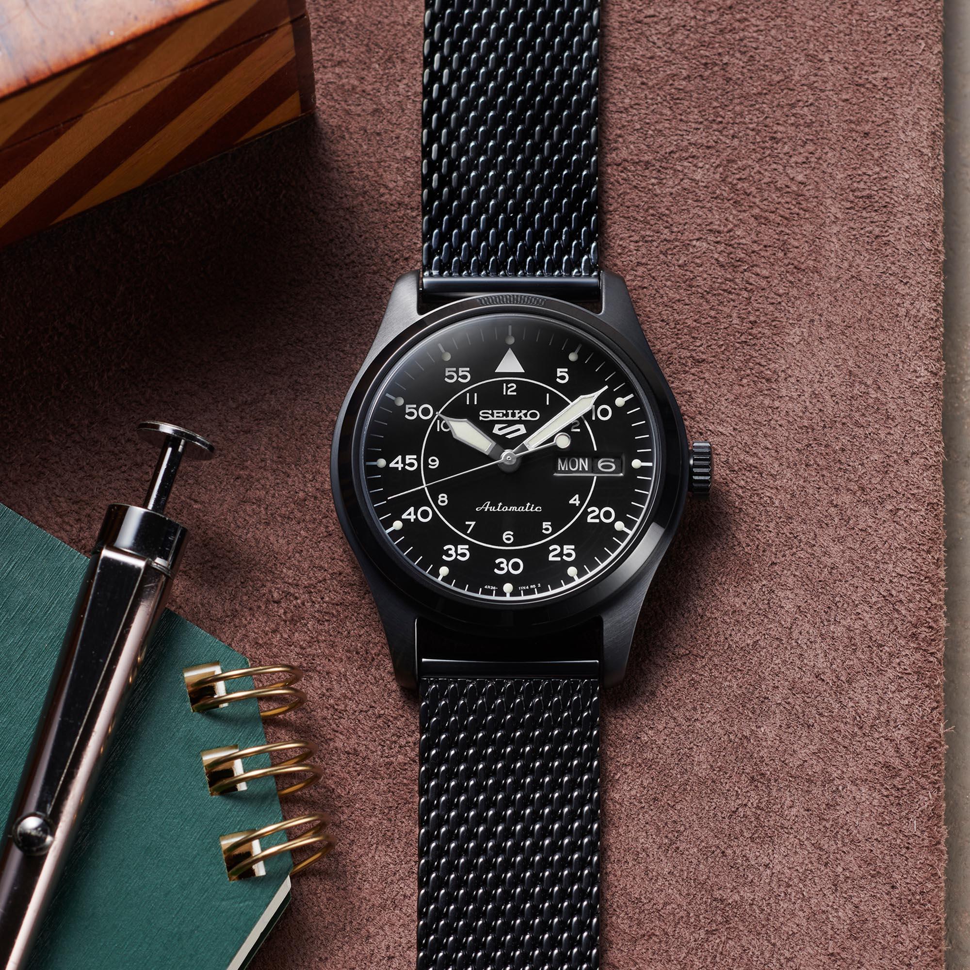 Black Seiko watch with mesh band on a textured surface with pen and notebook.
