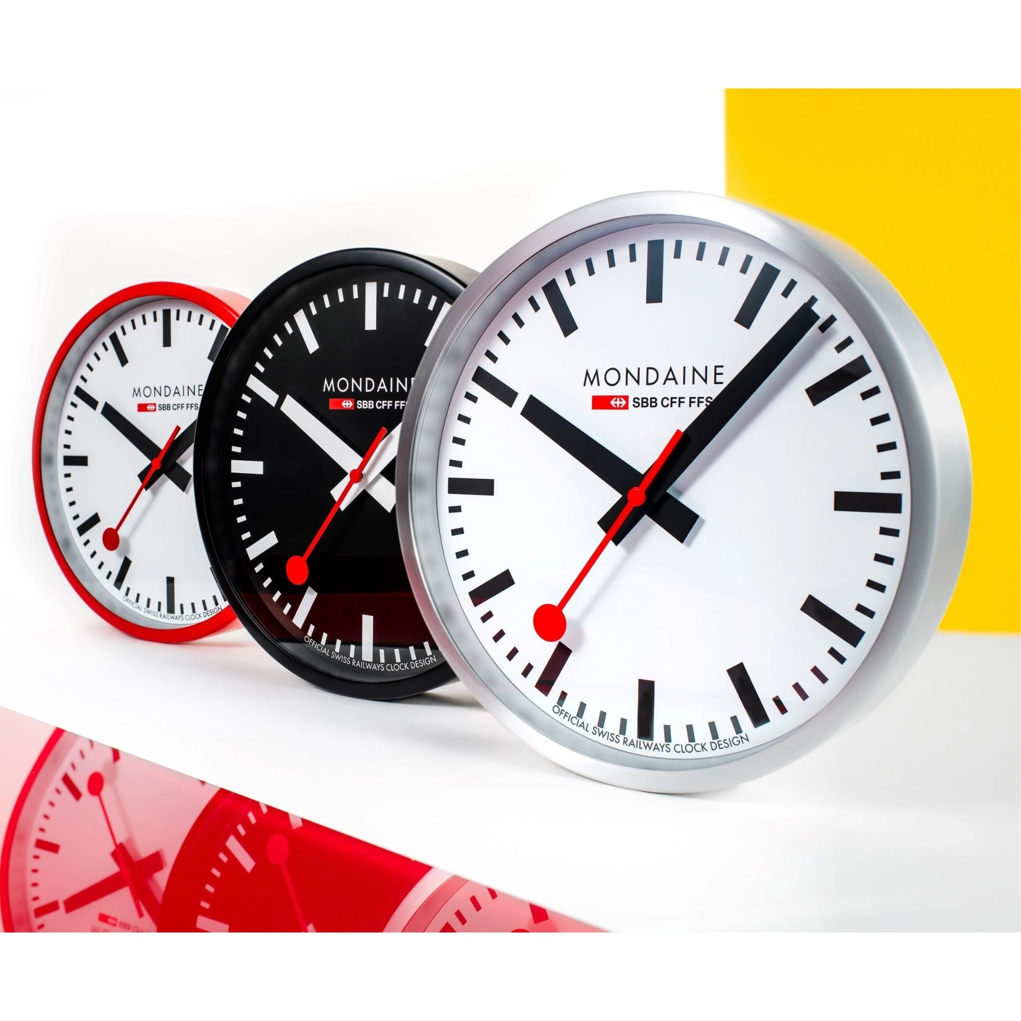 Three Mondaine wall clocks lined up, displaying red, black, and silver case finish options.