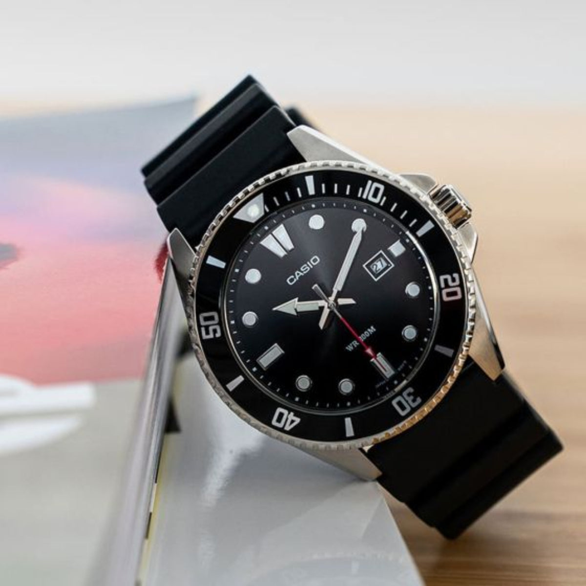 Casio Marlin MDV-107-1A1VEF resting against a magazine on a wooden surface next to a notebook