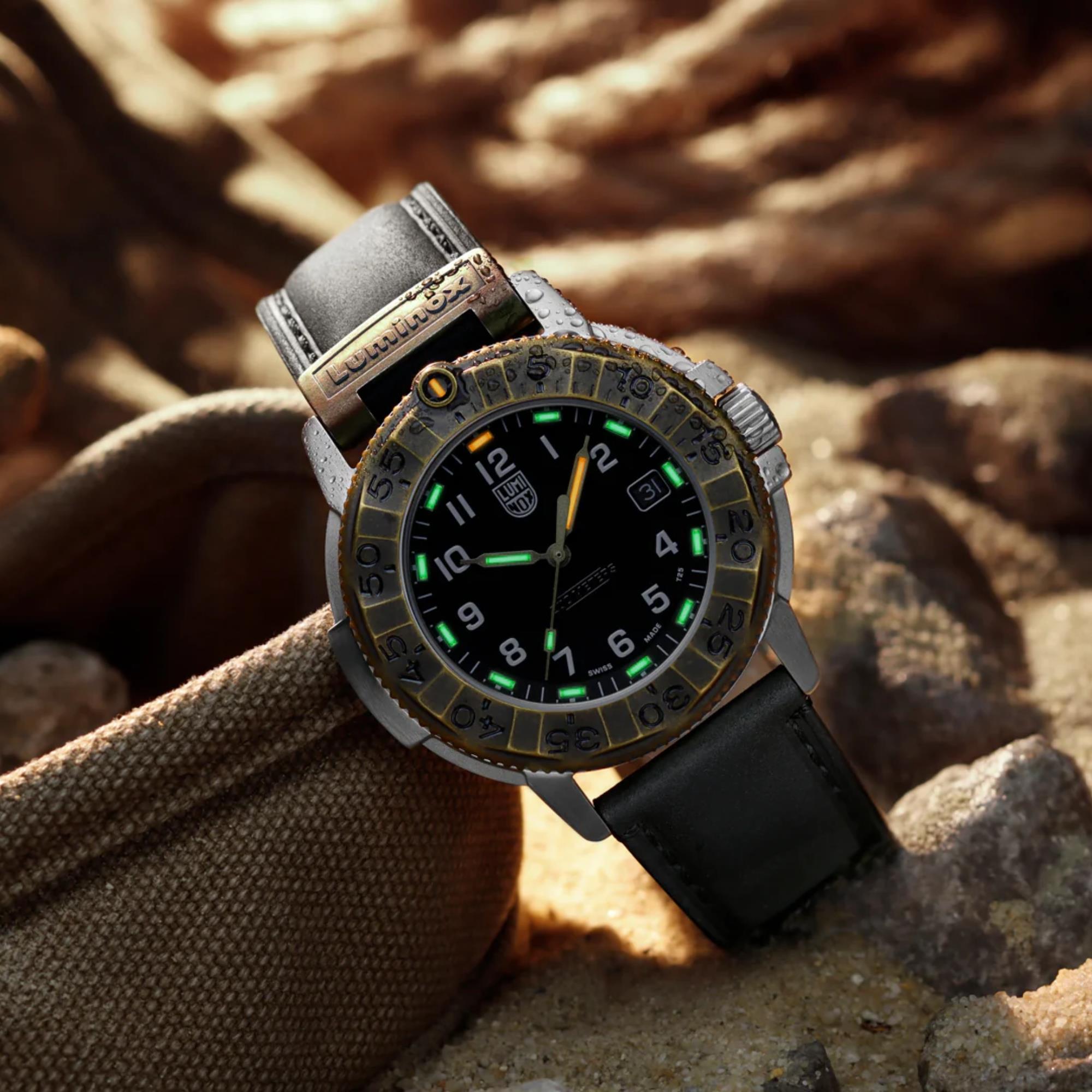 Close-up of the Luminox Adventure Watch XL.1301 resting on a canvas bag and stones, highlighting the glowing green and orange luminous markers on the black dial and its bronze bezel