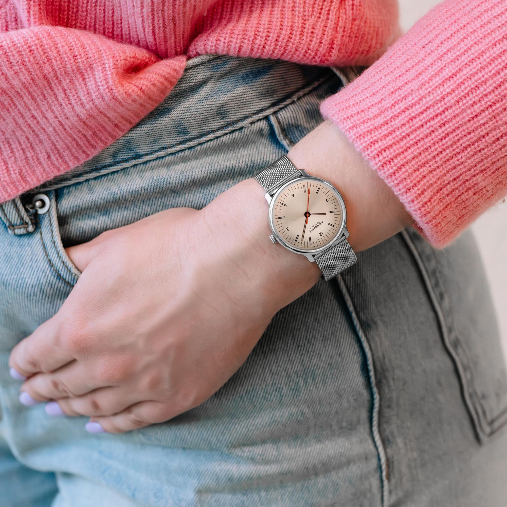 Mondaine watch worn on a wrist, paired with jeans and a pink sweater, highlighting its slim case and elegant profile in daily wear