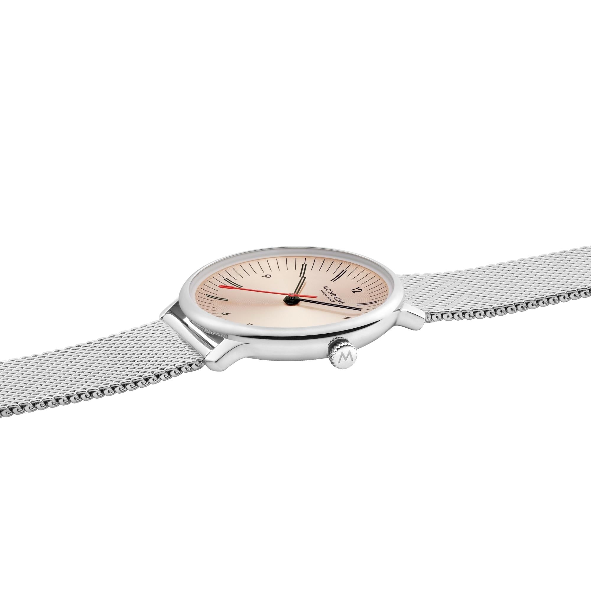 Angled side shot of the watch laying flat, displaying the textured winding crown stamped with the Mondaine "M" logo and the smooth transition between the case and mesh bracelet