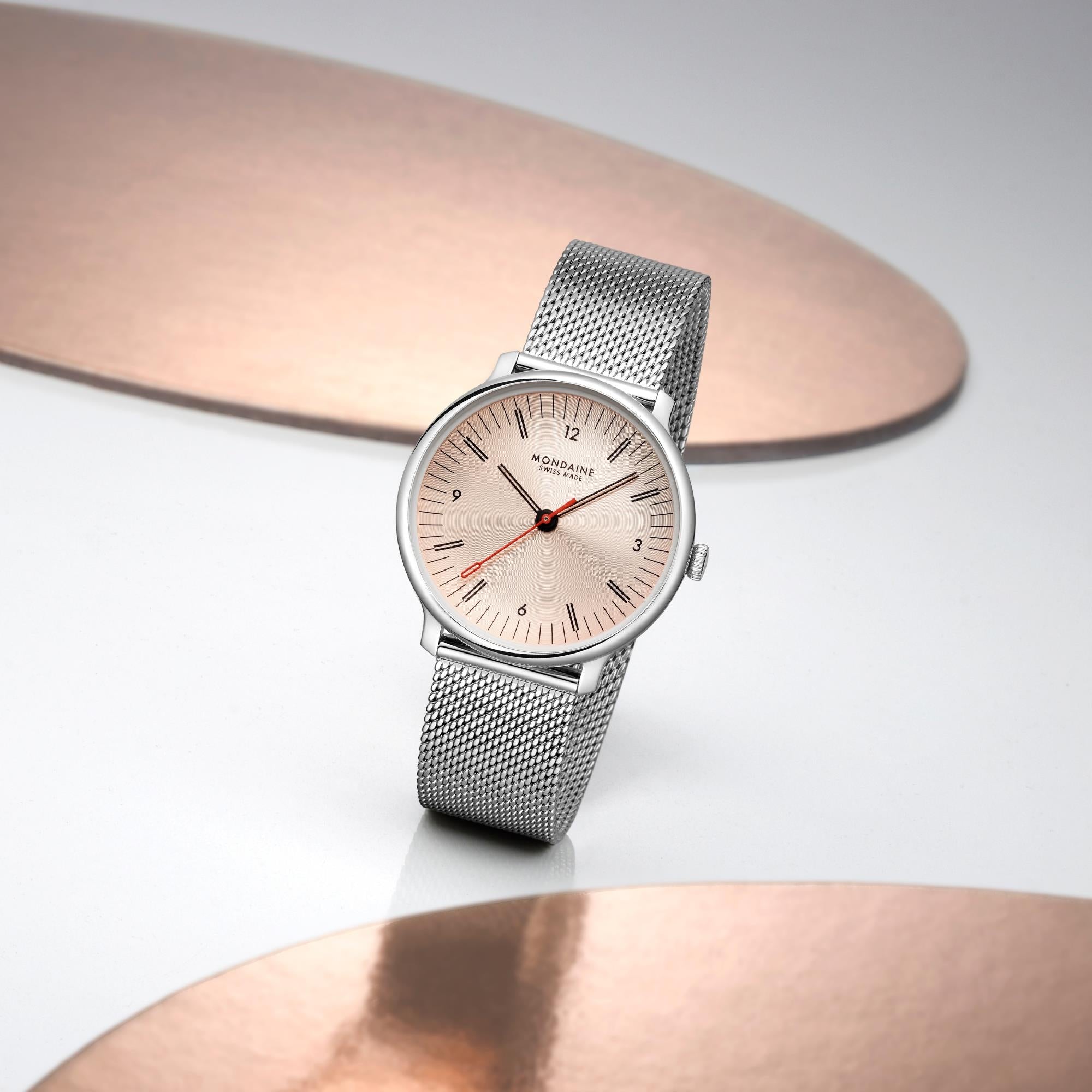 Mondaine watch standing upright on a white surface, framed by two decorative, curved copper or rose gold metallic accent pieces in the background